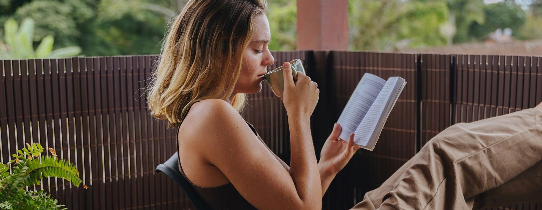 A woman sitting on a patio reading a book and sipping coffee.